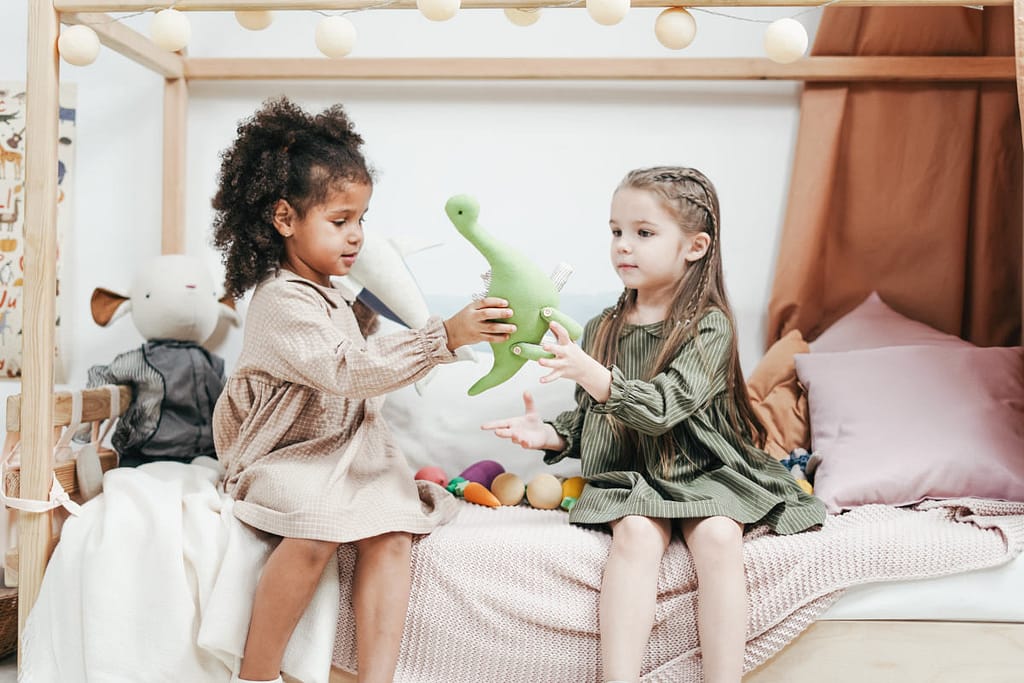 Two friends displaying the Fruit of the Spirit Goodness by sharing a stuffed animal while sitting on a bed together. 