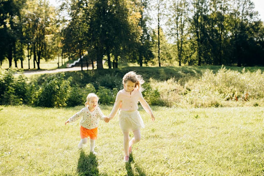 An older sibling holding her younger sibling's hand, walking through a grassy field, displaying the Fruit of the Spirit Kindness towards her younger sister.