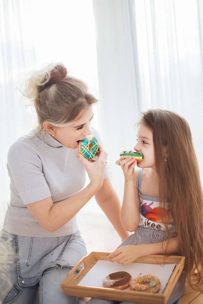 A mom and daughter smiling at each other while taking a bite out of a donut after they just enjoyed cleaning their kitchen